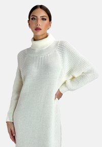 Hvid strikket sweaterkjole med høj hals, løs pasform og struktureret mønster. Har lange ærmer og sidelommer.