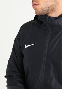 Nike sort vindjakke med hel lynlås, justerbar hætte og en hvid logoakcent på brystet, lavet af et glat, let stof.