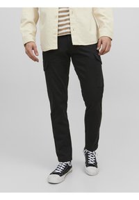 Svarta cargo-byxor med sidofickor och en smal design, tillsammans med svarta och vita sneakers, samt en beige skjorta med knappar över en randig topp.