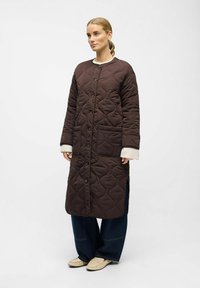 Femme portant un long manteau matelassé marron foncé avec de grandes poches, un pantalon large foncé, des poignets de pull crème et des mocassins beige clair.