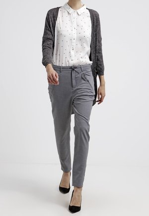 Vrouw loopt en draagt grijze broek met trekkoord, wit overhemd met kleine zwarte stippen, een grijze cardigan en zwarte hakken.