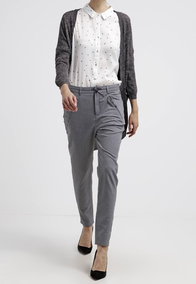 Femme marchant portant un pantalon gris à cordon, une chemise blanche à boutons avec de petits pois noirs, un cardigan gris et des talons noirs.