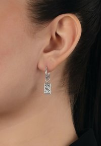 Ingrandimento di un orecchio umano che indossa un orecchino a cerchio in argento con un ciondolo ornamentale rettangolare appeso.