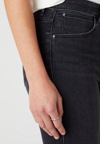 Vaqueros de mezclilla negros con un diseño ajustado, con costuras tonales, estilo de cinco bolsillos y una textura suave. Mano adornada con anillos de plata.