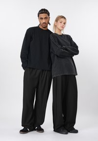 Δύο άτομα στέκονται δίπλα δίπλα. Φοράνε άνετα μαύρα ρούχα: μπλούζα με μακριά μανίκια, φαρδιά φούτερ και παντελόνια με φαρδιά μπατζάκια.