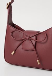 Burgundy faux leather handbag with a rounded shape, featuring a bow detail and gold-tone accents, smooth texture, and a shoulder strap.