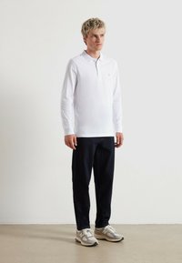 Polo bianco a maniche lunghe con un piccolo logo ricamato, abbinato a jeans scuri e sneaker grigie. Design semplice e casual.