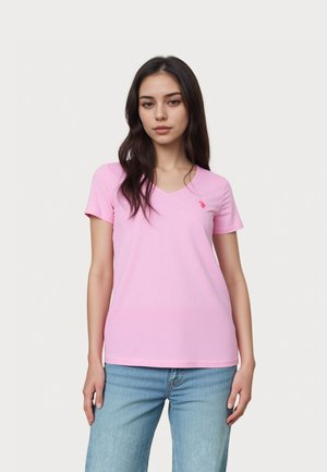 Mladá žena s dlouhými tmavými vlasy, která má na sobě světle růžové tričko s véčkovým výstřihem a světle modré džíny, stojí před jednobarevným bílým pozadím.