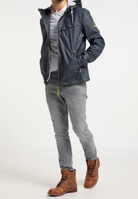 Homme portant une veste imperméable à capuche bleu marine, une chemise gris clair, un jean gris et des chaussures en cuir brun à lacets, les mains dans les poches de la veste.
