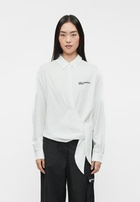 Camisa blanca de manga larga con cuello, que presenta un diseño cruzado y un lazo lateral. Logo negro en el lado izquierdo del pecho, textura suave. Combinada con pantalones negros.