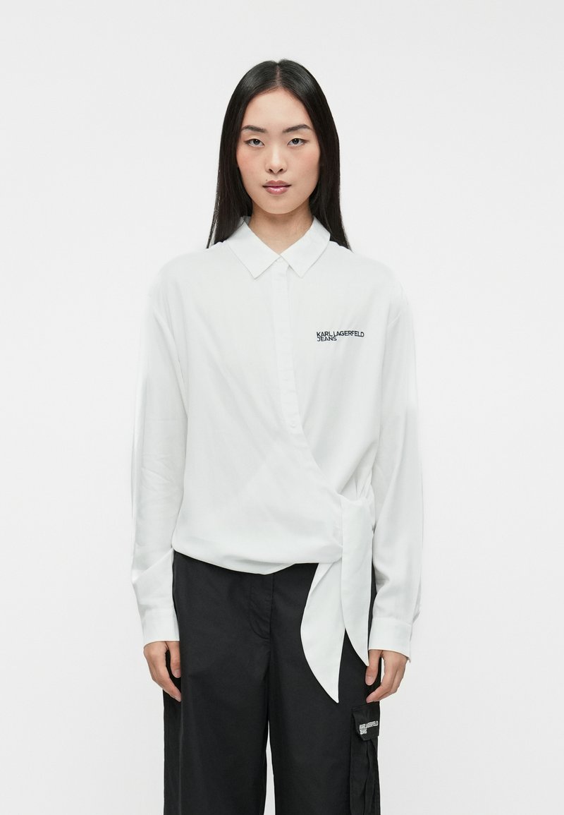 Camisa blanca de manga larga con cuello, que presenta un diseño cruzado y un lazo lateral. Logo negro en el lado izquierdo del pecho, textura suave. Combinada con pantalones negros.
