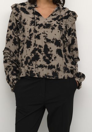 Vrouw draagt een taupekleurige blouse met lange mouwen, zwart abstract patroon en ruches, gecombineerd met een zwarte broek en handen in de zakken.