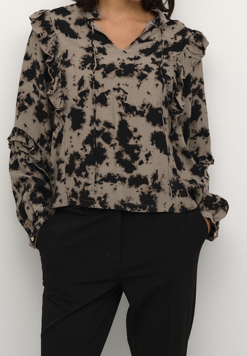 Femme portant un chemisier taupe à manches longues avec motif abstrait noir et détails à volants, associé à un pantalon noir et les mains dans les poches.