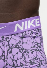 Μέση και άνω μέρος γοφού ατόμου που φοράει μοβ και μαύρα εμπριμέ εσώρουχα Nike με πλατύ μοβ ζωνάκι και μεγάλο λευκό λογότυπο NIKE.