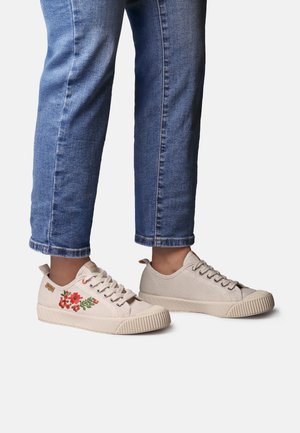 Beżowe sneakersy z kwiatową haftowaną, zaokrąglonym noskiem i teksturowaną gumową podeszwą, zestawione z krótkimi niebieskimi dżinsami.