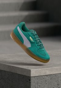 Vihreät mokkakengät, joissa on valkoinen PUMA-raita ja kultainen logo. Kumipohja ja tekstuurilliset nauhat viimeistelevät muotoilun.