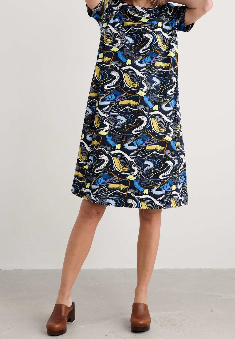 Femme portant une robe à manches courtes, longueur genou, avec des motifs abstraits bleus, jaunes, blancs et noirs, associée à des sabots bruns à talons.