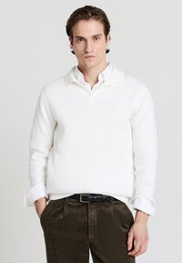 Maglione bianco a coste con colletto, abbinato a pantaloni in velluto a coste di colore verde scuro. Tessuto leggero, vestibilità casual e dettaglio dei polsini visibile.