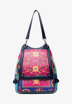 Kobieta ubrana w jasnoniebieskie dżinsy, czarny prążkowany sweter, białe trampki i kolorowy kwiatowy plecak, stojąca na tle białego tła.