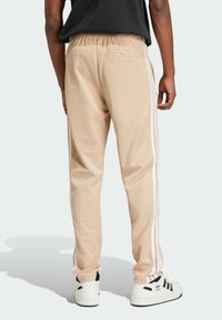 Pantalones deportivos beiges con rayas blancas en los laterales, cinturilla elástica y bolsillo trasero. Combinados con zapatillas negras y blancas. Textura de tela suave.