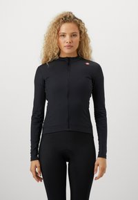 Kvinne med krøllete blondt hår iført en tettsittende svart langermet sykkeltrøye og svarte sykkelshorts, som står mot en ensfarget hvit bakgrunn.