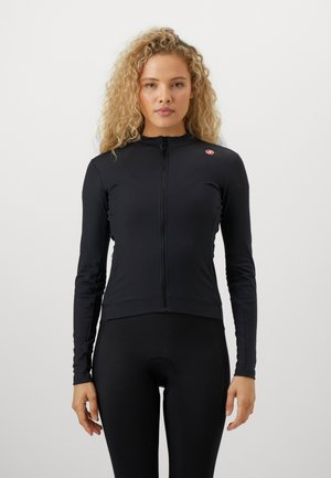 Frau mit lockigem blonden Haar, die ein eng anliegendes schwarzes Langarm-Radtrikots und schwarze Fahrradhosen trägt, steht vor einem schlichten weißen Hintergrund.
