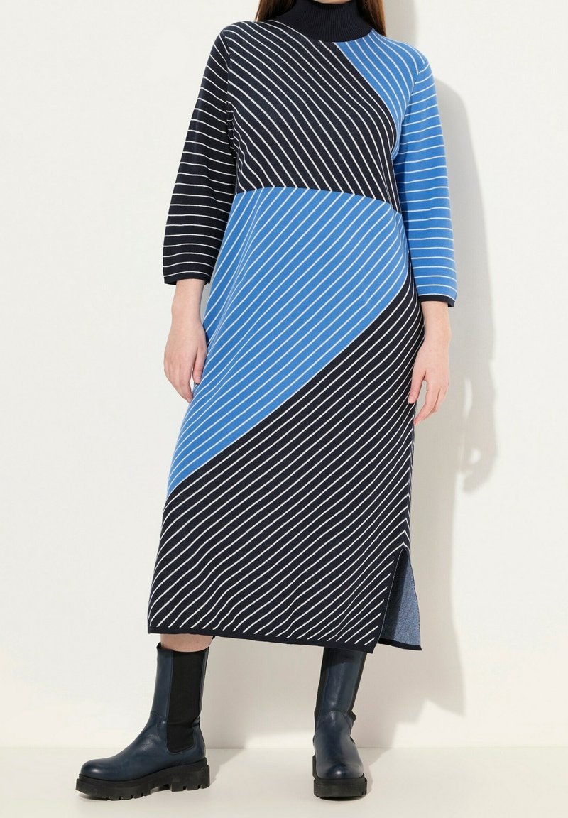 Femme portant une robe longue en maille noire et bleue à rayures, manches trois-quarts, et des bottines noires à semelles épaisses.