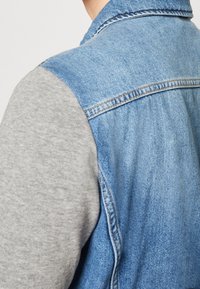 Jeansväst med grå sweatshirtärmar; har en krage och en mittbakssöm; textil med textur i blå och grå kontrasterande färger.