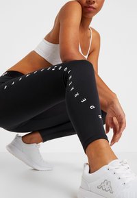 Black athletic leggings with a tight fit, featuring white text along the leg. Paired with a white sports bra and white sneakers with mesh texture.