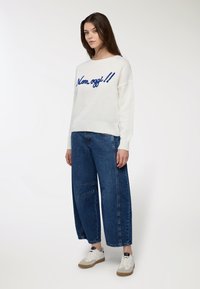 Maglione bianco lavorato a maglia con testo ricamato blu "Non oggi!!", abbinato a jeans in denim blu a gamba larga e sneaker bianche.