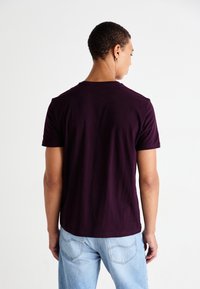 T-shirt à manches courtes, col rond, en coton violet foncé. Texture lisse et design simple. Porté avec un jean bleu clair.