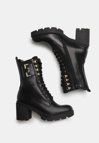 Schwarze Lederstiefeletten mit einem klobigen Absatz, goldenen Beschlägen, Schnürvorderseite und seitlichem Reißverschluss. Texturiertes Sohlenprofil mit robustem Design.