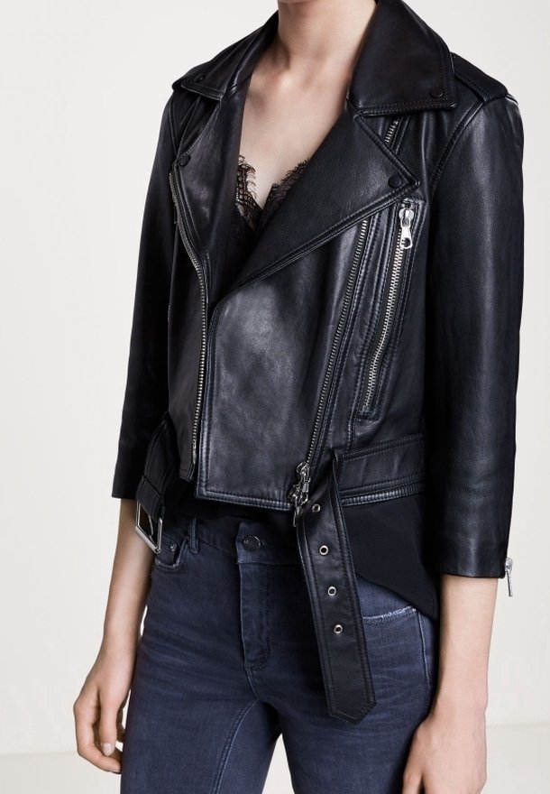 Femme portant une veste courte en cuir noir de motard avec des fermetures éclair argentées, une taille ceinturée et un jean bleu foncé, sur un fond clair uni.