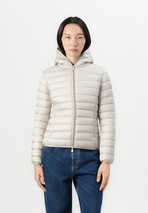 Licht beige pufferjack met horizontale stiksels, een ritssluiting aan de voorkant en een capuchon. Beschikt over strakke mouwen en zijzakken. Draag het over blauwe jeans.