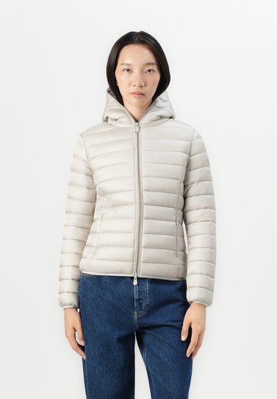 Doudoune beige clair avec un matelassage horizontal, une fermeture éclair à l'avant et une capuche. Comprend des poignets ajustés et des poches latérales. Portée sur un jean bleu.