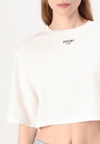 Balta trumpa medvilninė marškinėlių palaidinė su apvaliu kaklu ir trumpomis rankovėmis. Centre yra juodas „JOOP! JEANS“ logotipas.