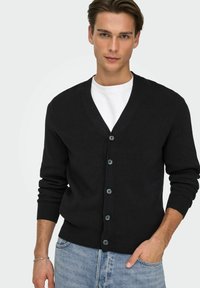 Cardigan nero in tessuto lavorato a maglia con scollo a V, dotato di sei bottoni scuri, polsini a coste e vestibilità comoda. Indossato sopra una t-shirt bianca.