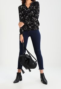 Blusa nera a fiori con maniche lunghe, jeans skinny blu scuro, stivaletti neri alla caviglia e una borsa in pelle nera con frange.