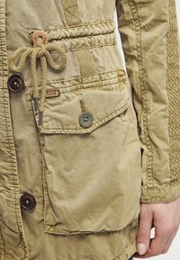 Veste kaki avec une manche texturée, des accents de boutons, une taille ajustable avec cordon de serrage, et une poche avant avec rabat et fermeture par bouton.