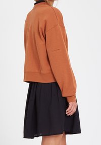 Sweat-shirt marron à coupe décontractée, avec poignets et col côtelés, associé à une jupe noire. Détail logo sur la manche gauche.