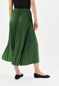 Zielona plisowana spódnica z elastycznym pasem, zwiewny materiał i gładka tekstura, zestawiona z czarnymi płaskimi butami z akcentem w postaci kokardki.