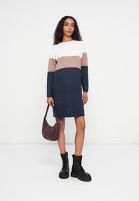 Abito in maglia con tre blocchi di colore orizzontali: crema, marrone chiaro e blu navy. Maniche lunghe, scollo rotondo e vestibilità rilassata. Stivali neri con lacci e borsa bordeaux.