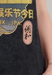 Top negro sin mangas con texto impreso en oro y plata, un parche texturizado en bronce con caracteres, y un acabado de tela suave y lisa.