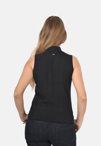 Donna con lunghi capelli castani chiari, che indossa un top nero senza maniche e jeans scuri, in piedi con le mani sui fianchi, rivolta lontano dalla macchina fotografica.