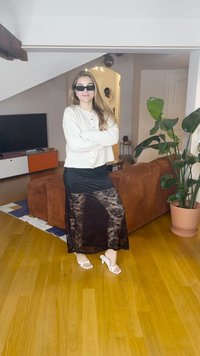 Hvit knappet cardigan, svart blondeskjørt, hvite åpne tåhæl, og svarte solbriller, foran et minimalistisk interiør med planter.