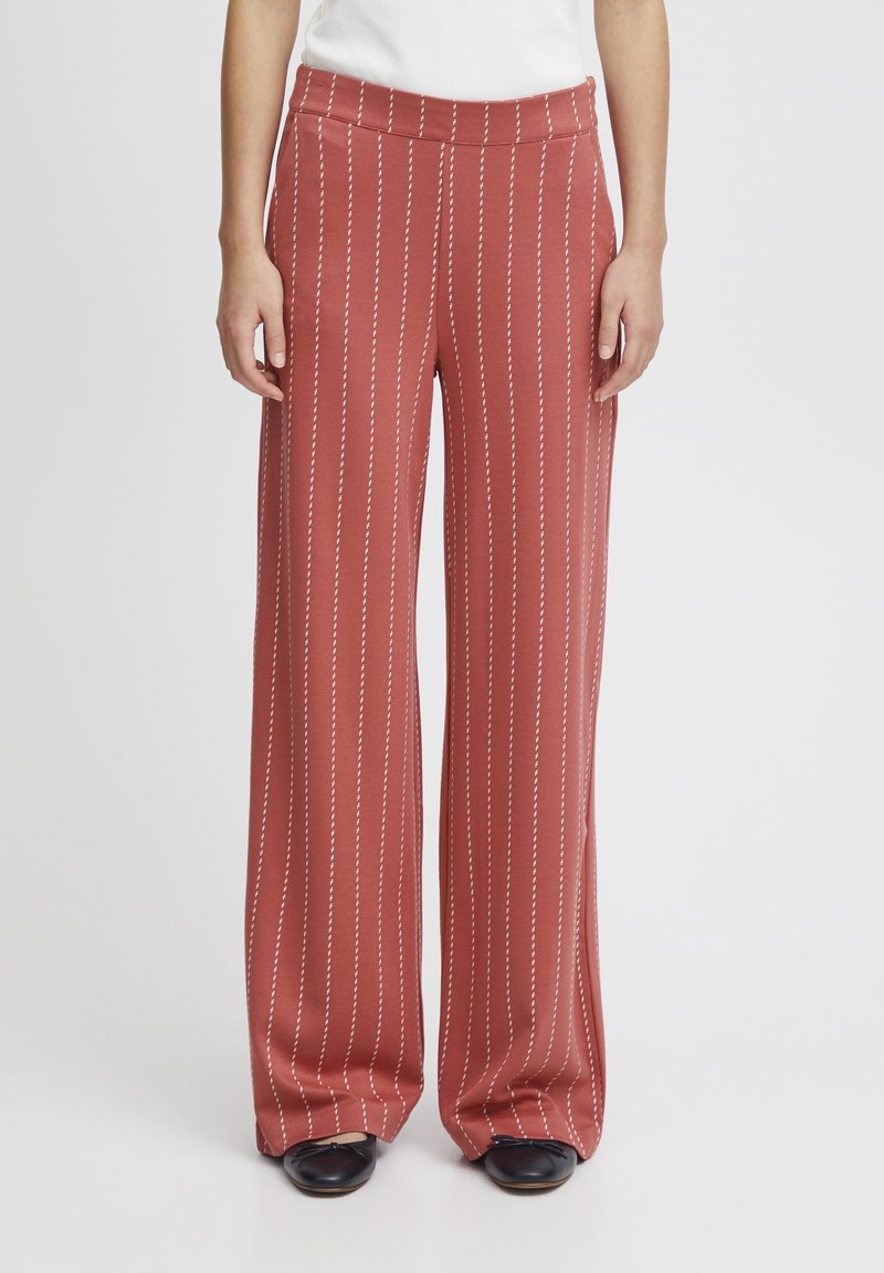 Femme portant un pantalon large couleur rouille avec des rayures verticales blanches et des chaussures noires, debout devant un fond uni.