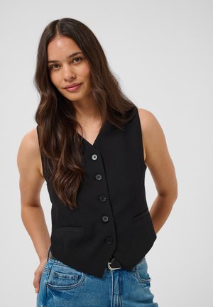 Femme aux longs cheveux bruns portant un gilet noir boutonné et un jean bleu, debout les mains dans le dos devant un fond uni.