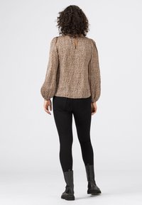 Langärmlige Bluse in Beige mit einem strukturierten Muster, ausgestattet mit einem Schlüssellochausschnitt hinten. Kombiniert mit schwarzen Leggings und schwarzen Stiefeletten.