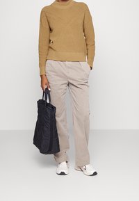 Maglione khaki a coste con motivo geometrico, pantaloni beige a gamba larga e una borsa a tracolla nera texturizzata. Sneakers bianche con dettagli colorati.