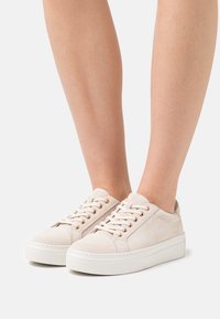 Světle béžové semišové tenisky s kulatou špičkou, bílou platformovou podrážkou, zlatými kovovými očky a plochými tkaničkami.